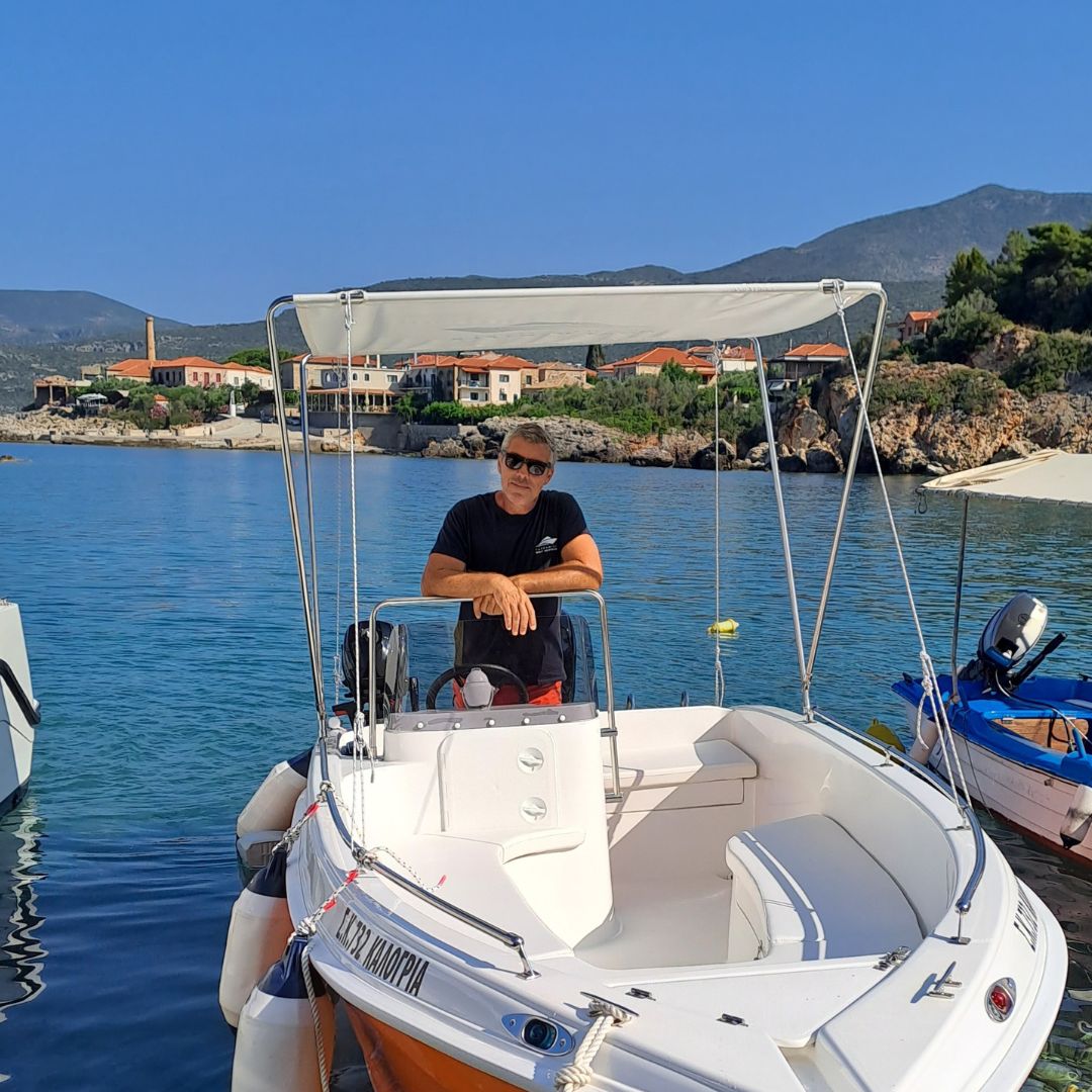 This is Christos, one of our tea members, looking at the camera while standing inside the "Kalogria" boat, at Kardamili Boat Harbor. In the background there are some houses of Kardamili village. Christos is smiling.