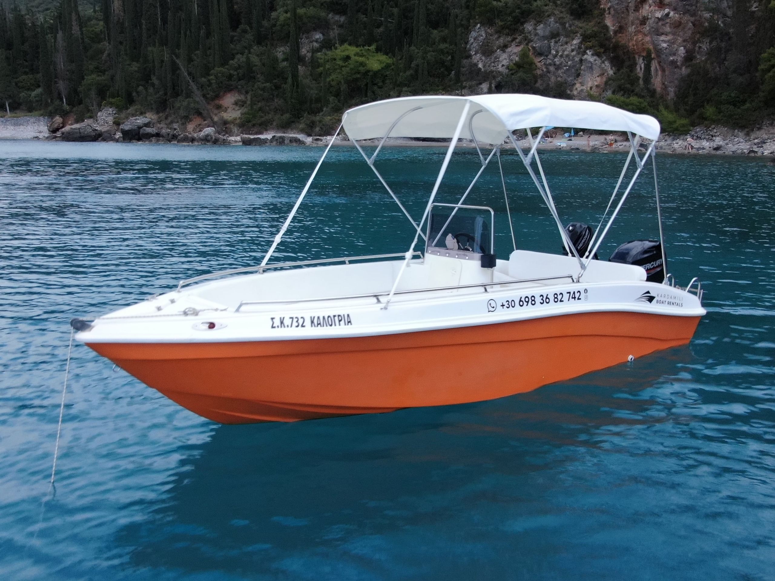 This is an image of our orange Kardamili Boat Rentals Boat, called Kalogria, at the Mani sea. At the background there is a coast with trees and rocks.