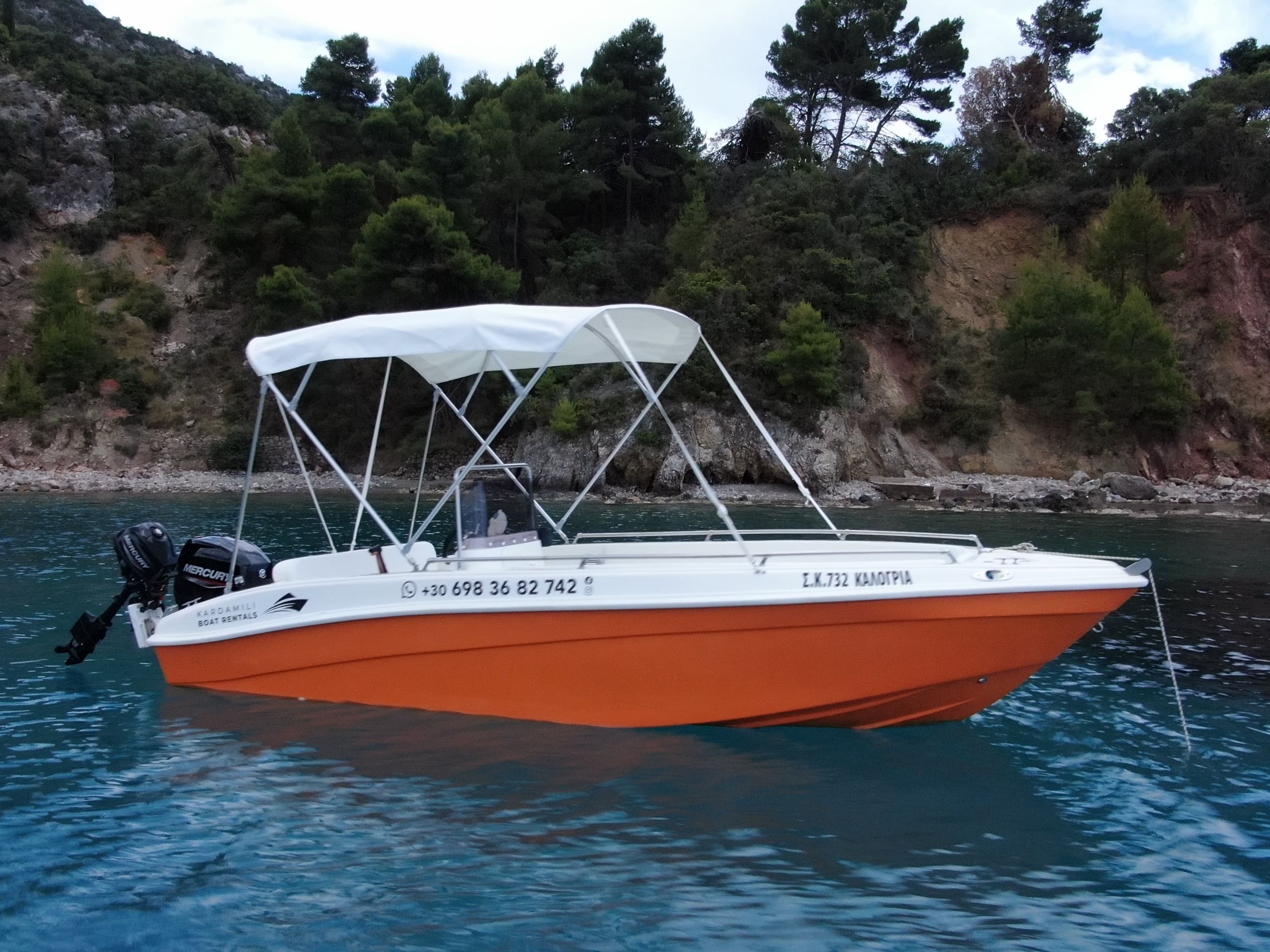 This is an image of our orange Kardamili Boat Rentals Boat, called Kalogria, at the Mani sea. At the background there is a coast with trees and rocks.