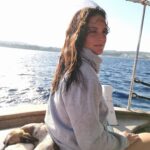 This is Sofia Kyriakoulakou. She is sitting in a boat in the middle of the sea. She is wearing a grey sweatshirt and behind her there is a little dog sleeping. She has brown hair and is of white color. She is smiling,