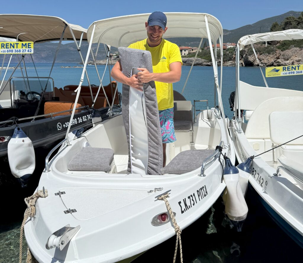 This is an image of Vasilis wearing shorts and a Kardamili Boat Rentals sweater, standing in the Kalamitsi Boat at Kardamili harbor, and holding a pillow with a pillow cover.