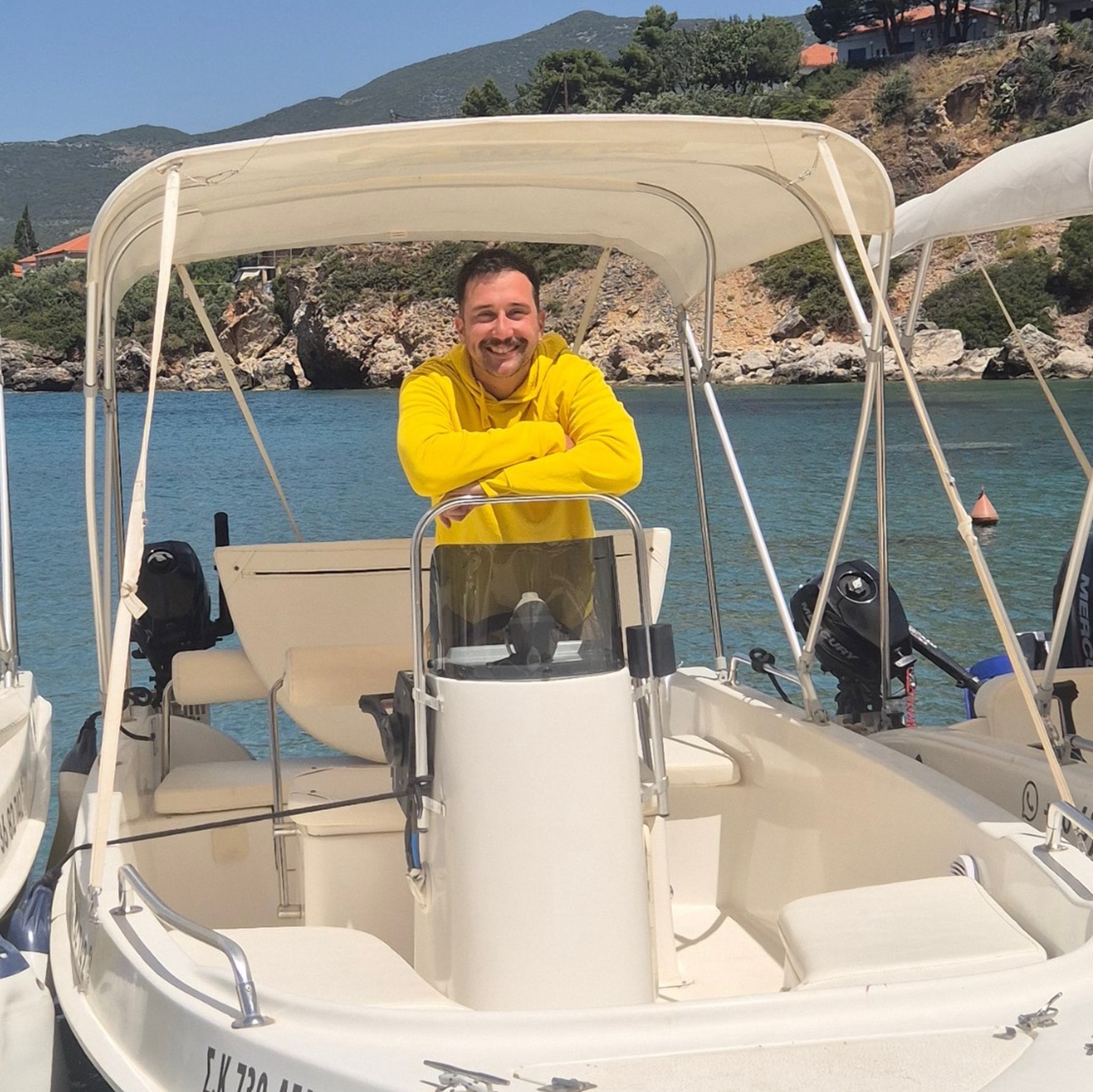 This is Kostas Rebholz. He is standing in a boatv of Kardamili Boat Rentals. The blue boat named Delfinia. Behind him is Kardamili harbor. He is wearing a yellow Kardamili Boat Rentals tshirt. He is smiling.